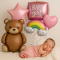 Baby girl celebration setup with large teddy bear balloon, pink star and heart foil balloons, rainbow with smiling clouds, and a newborn baby wrapped in pink — perfect for baby shower or photo backdrop.